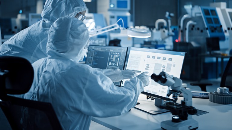 Zwei Personen in Schutzanzügen in Laborumgebung interagieren und zeigen auf einen Bildschirm.